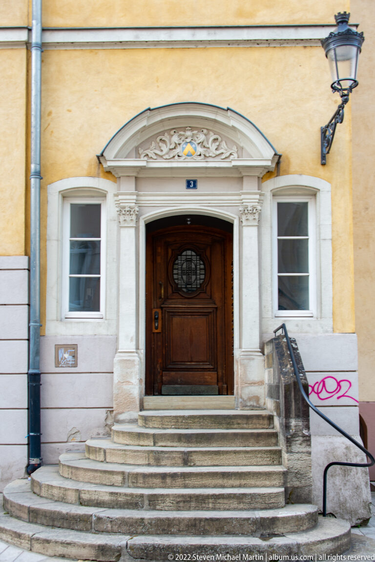 Dark wooden doorway with oval glazing reached by broad curved stone steps beneath a classical surround and wall lantern.