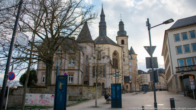 Historic church with a pointed spire, traceried window, and adjoining tower rises along a narrow Luxembourg City street.