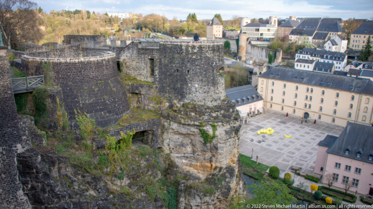 Massive stone fortification ruins and rock-cut openings overlook a paved lower courtyard and nearby buildings.