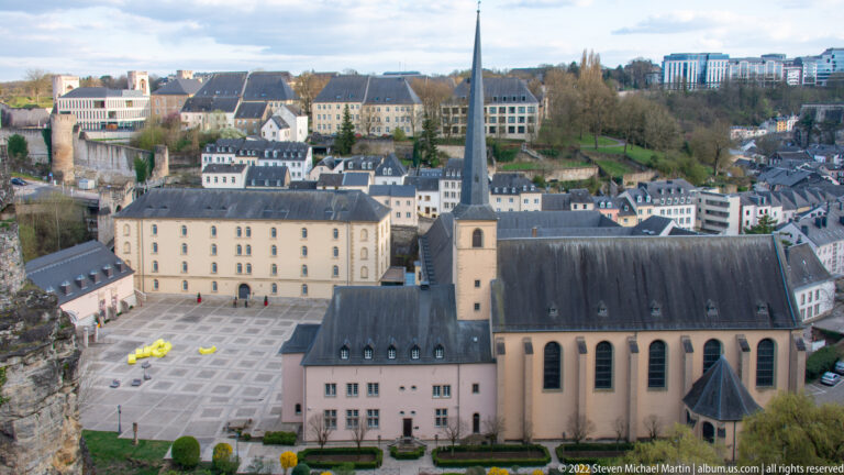 Long slate-roofed church with a tall square tower and slender spire stands among lower-city buildings below the plateau.
