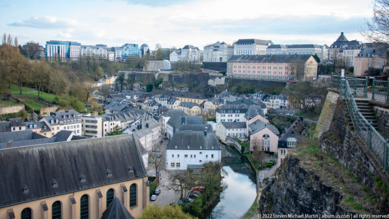Historic lower-city houses cluster along a river beneath steep fortified cliffs and the upper city.