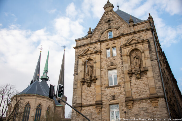 Ornate historic stone façade stands beside a church apse and several pointed spires under a bright sky.