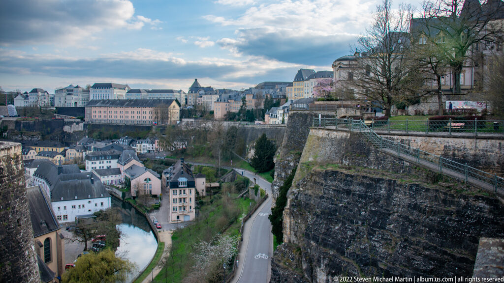 High fortified escarpment overlooks a river, roadway, lower-town houses, and upper-city buildings across the valley.