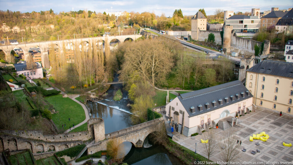 Stone bridge crosses a narrow river below landscaped paths, fortified walls, and upper-city buildings.