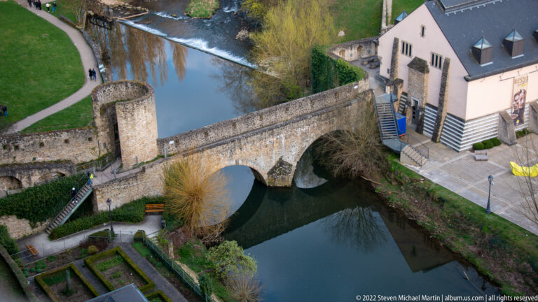 Single-arch stone bridge spans a calm river beside a pale historic building, tower, paths, and gardens.