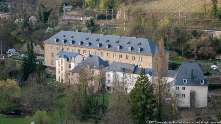 Large historic building complex with steep slate roofs and dormers sits on a wooded valley terrace.
