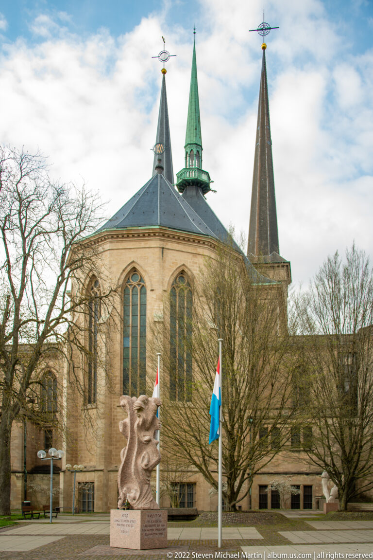 Polygonal church apse with tall windows and three slender spires behind flags and a stone sculpture.