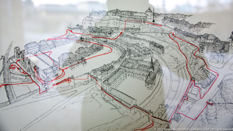 Axonometric map of Luxembourg's historic fortifications with a red route traced through walls, bastions, and streets.