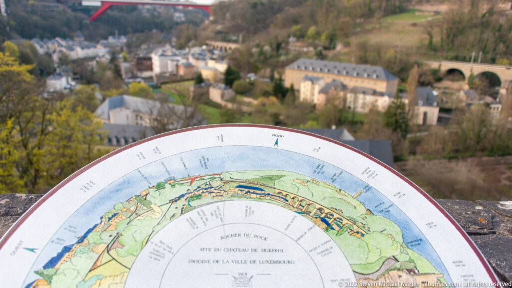 Semicircular painted orientation panel overlooks Luxembourg's valley, historic buildings, and an arched bridge.
