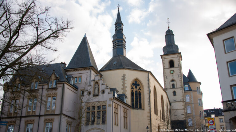 Historic church with tall pointed window, steep roof, clock tower, and nearby turrets rises above adjoining buildings.