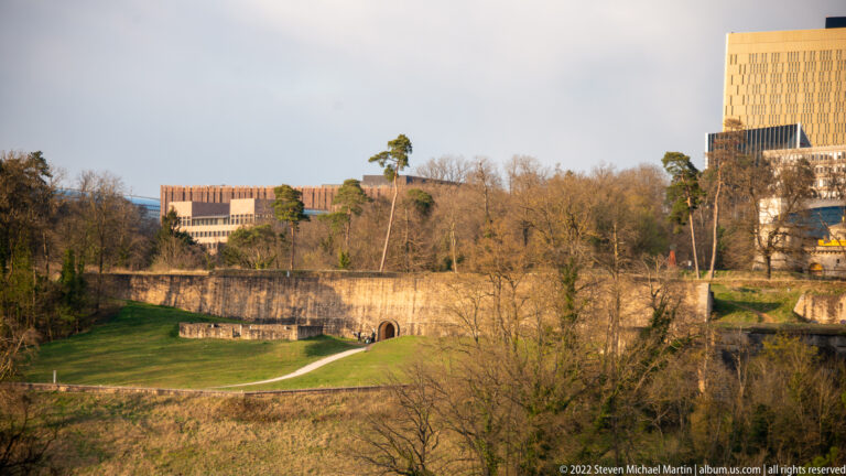Grassy defensive landscape with stone wall and arched opening below trees and modern upper-city buildings.