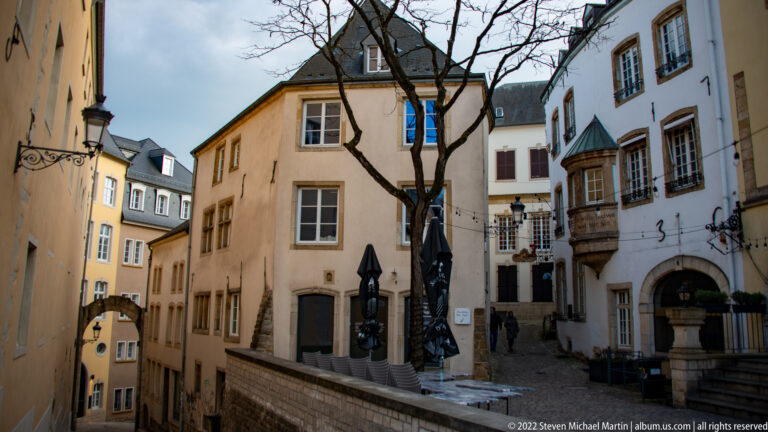 Narrow historic lane with pastel façades, café tables, lanterns, and a projecting bay window.
