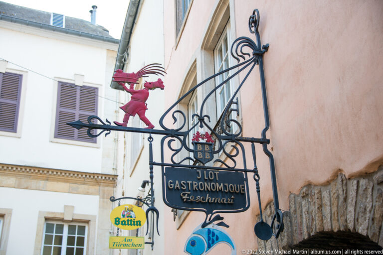 Decorative wrought-iron restaurant sign with red figure, shields, fish motif, and 'Gastromonique' lettering.