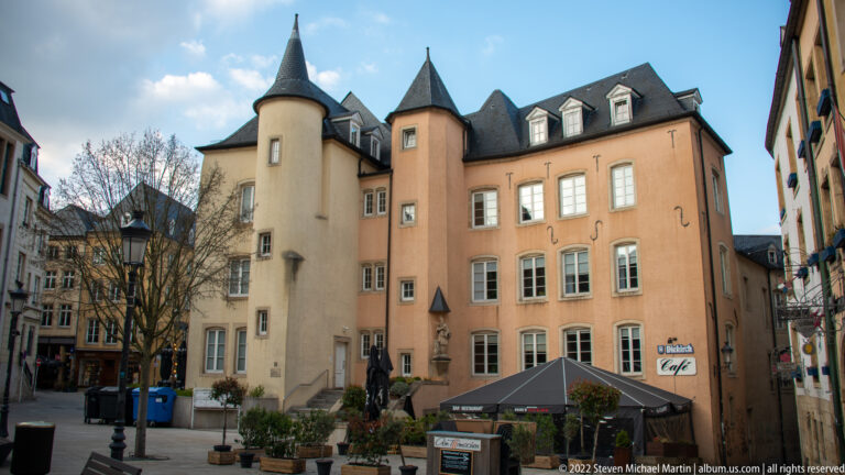 Pastel historic buildings with two conical-roofed corner turrets overlook café seating in a small courtyard.
