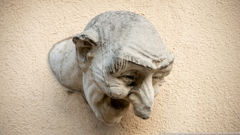 Weathered stone grotesque head with prominent nose and open mouth projects from a plain stucco wall.