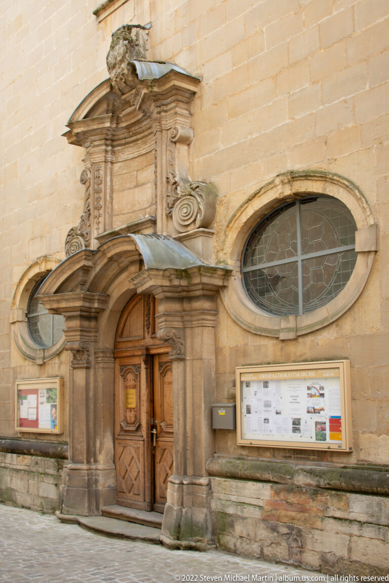Ornate carved stone church doorway with pilasters and curved pediment beside a large oval window.