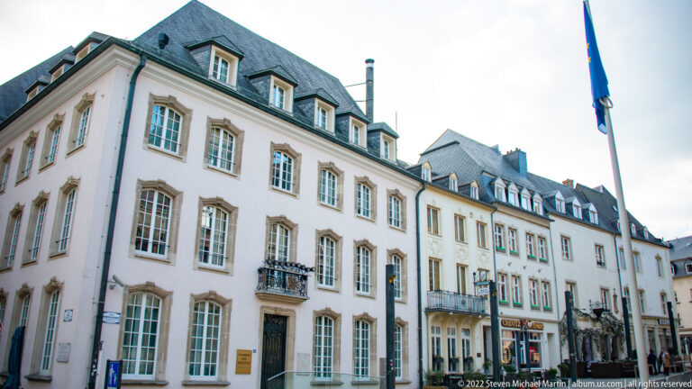 Row of pale historic townhouses with tall windows, iron balconies, slate roofs, dormers, and ground-floor shops.