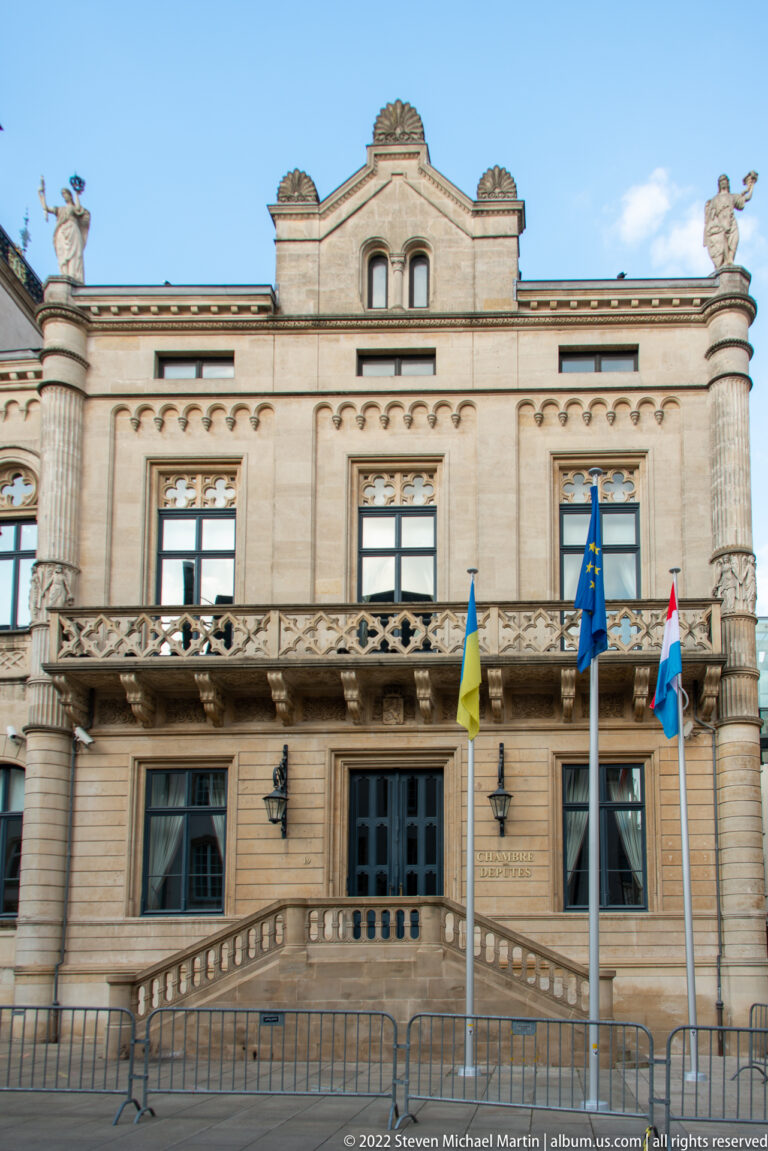 Symmetrical stone civic building with broad entrance stair, balustrades, roof sculptures, and official flags.