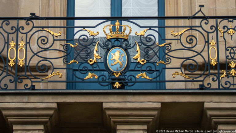 Blue shield with crowned golden lion and gilded crown set within ornate black-and-gold palace balcony ironwork.