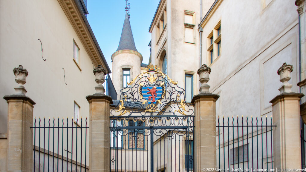 Ornamental palace gate with crowned Luxembourg lion coat of arms, gilded scrollwork, stone posts, and turret beyond.