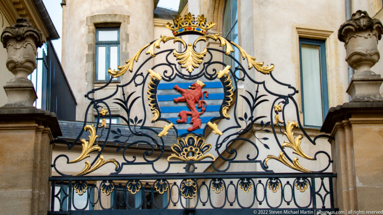 Red lion rampant on blue-and-white striped shield beneath a jeweled crown within gilded wrought-iron gate ornament.
