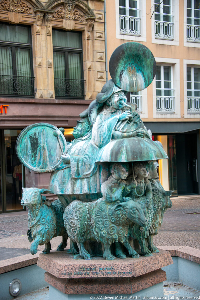 Large green-patinated bronze fountain sculpture with layered organic forms stands in a shallow urban basin.