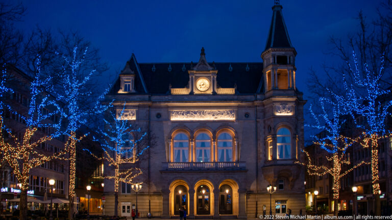 Cercle Cité illuminated at night with central clock, arched entrances, corner tower, and blue-lit trees.