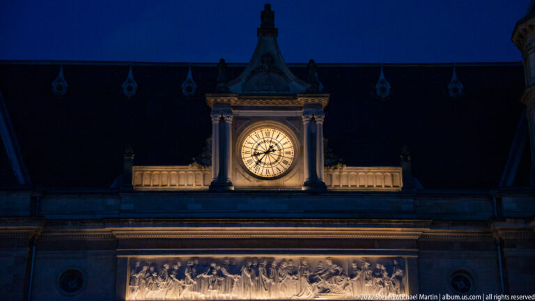 Illuminated round clock and long sculpted frieze on the central façade of Cercle Cité at night.