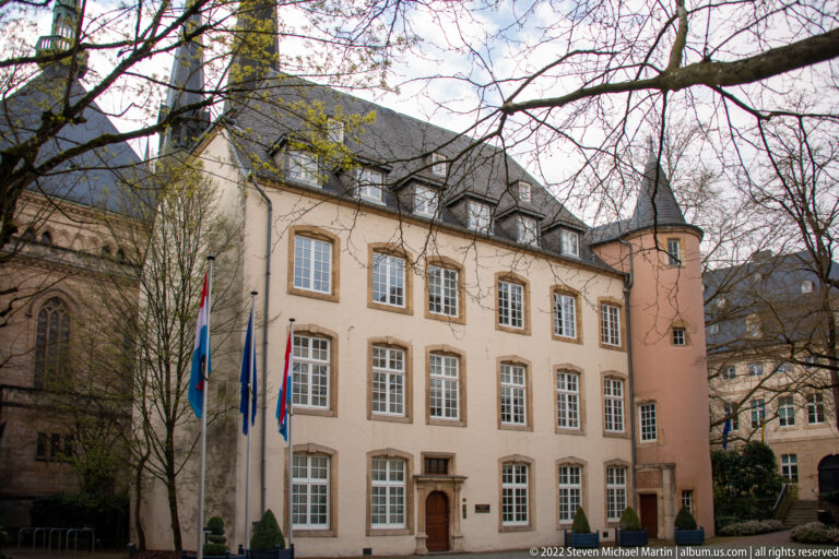 Three-story historic government building with rows of tall windows, flagpoles, clipped shrubs, and steep slate roofs.