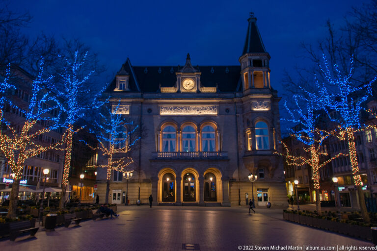 Wide night view across Place d'Armes toward illuminated Cercle Cité framed by blue- and gold-lit trees.