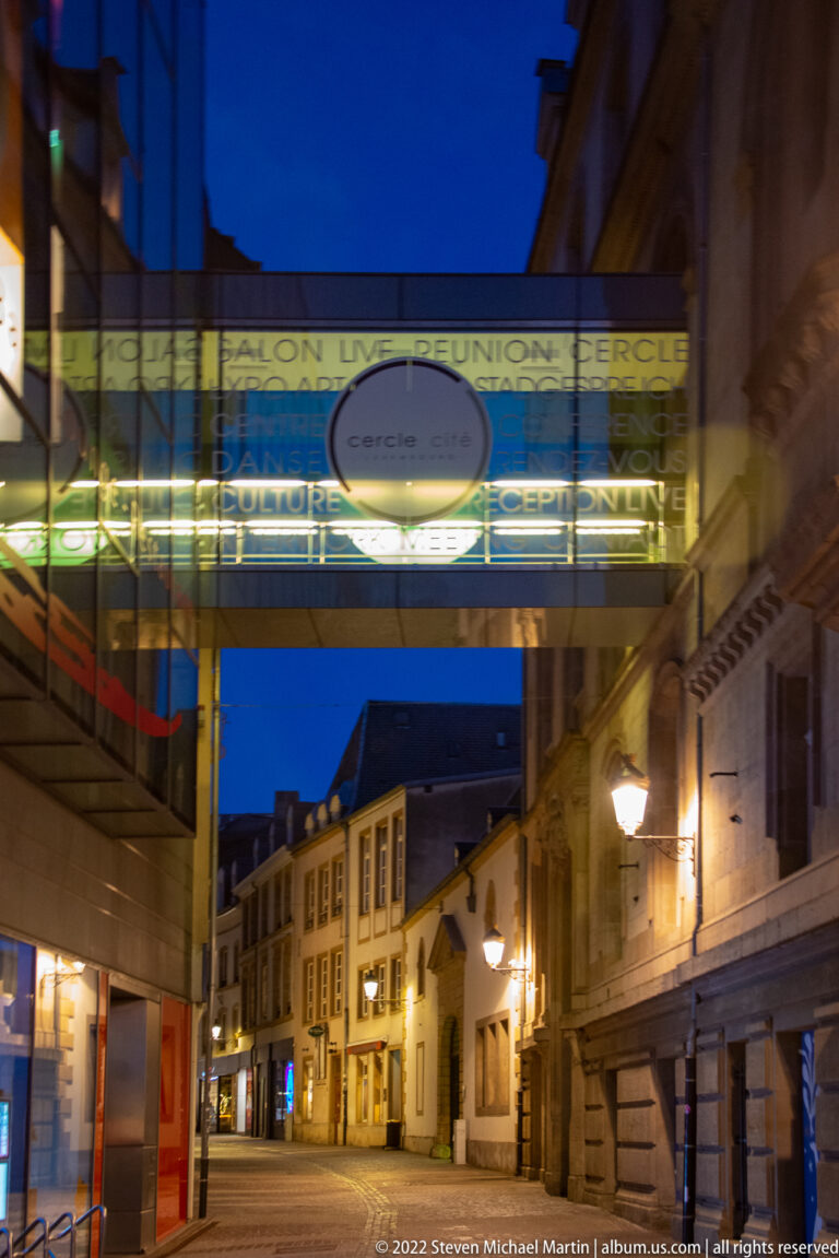 Glass pedestrian bridge marked Cercle Cité spans a narrow, lamp-lit historic street at night.