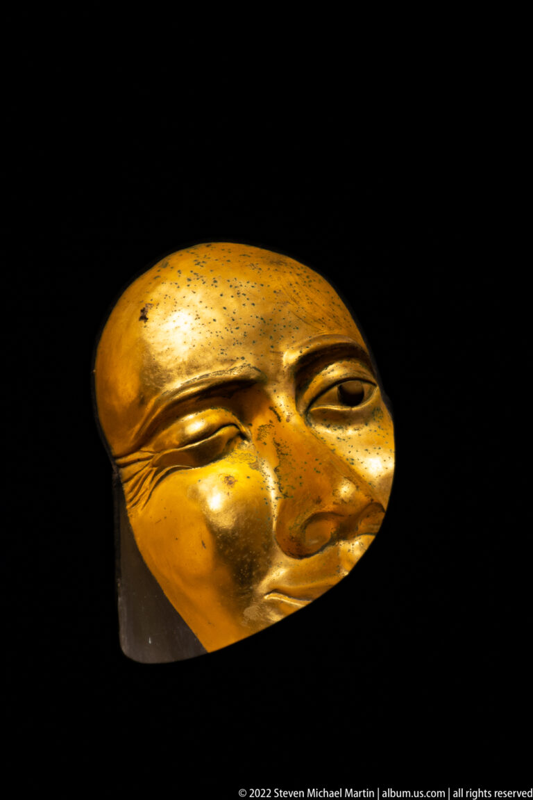Gilded stylized human face mask with almond-shaped eyes and broad forehead displayed against black.