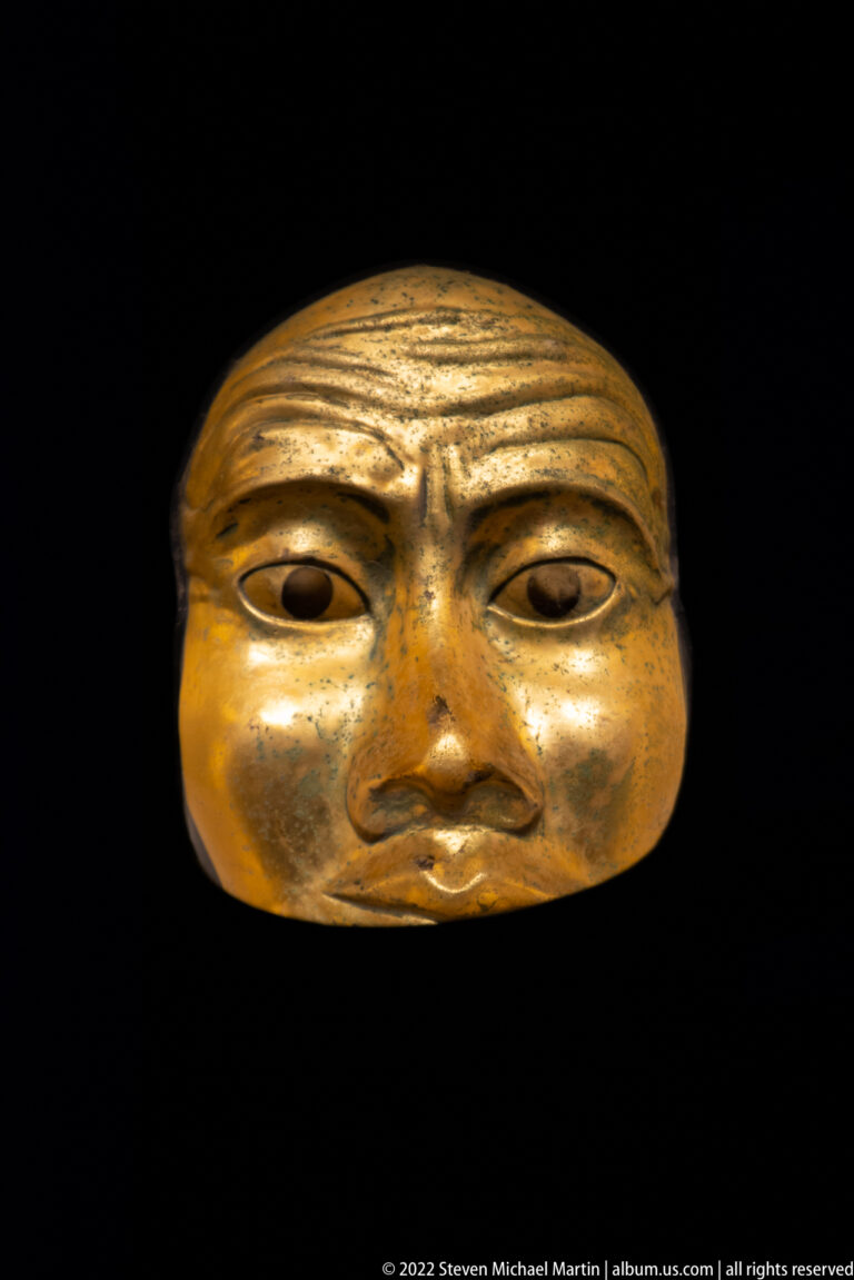 Frontal gilded face mask with heavy brow, almond-shaped eyes, broad nose, and worn metallic surface.