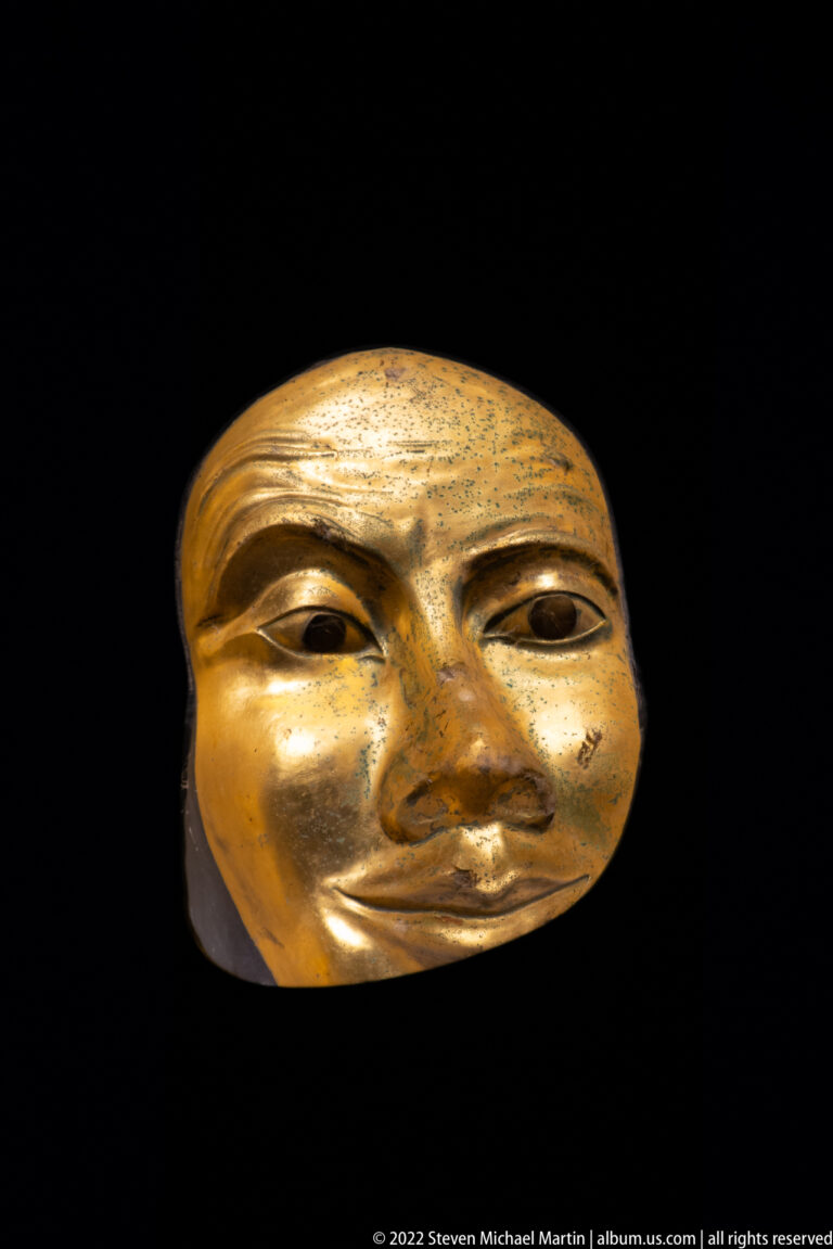 Rounded gilded face mask with asymmetrical eyes, broad nose, softly smiling lips, and mottled surface wear.