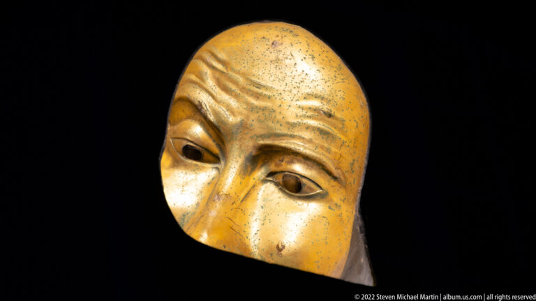 Elongated gilded face mask at a slight angle with narrow eyes, strong nose, brow creases, and spotted surface.