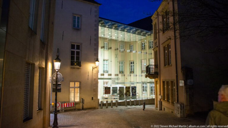 Narrow cobbled lane between historic façades leads toward a brightly illuminated modern courtyard building at night.