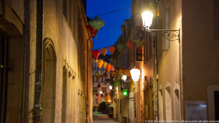 Narrow historic lane at night with colorful parasol-like decorations hanging overhead and warm street lamps below.