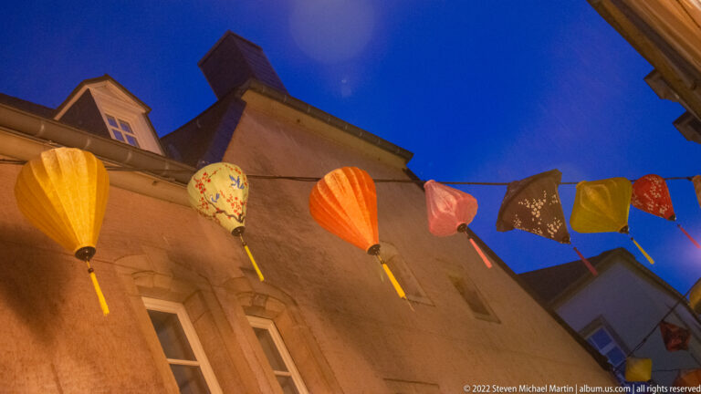 Colorful parasol-shaped street decorations hang above historic façades against a deep blue evening sky.