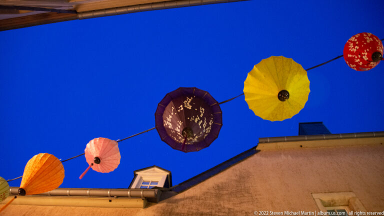 Upward view of colorful parasol-shaped ornaments suspended in a line against a deep blue sky.