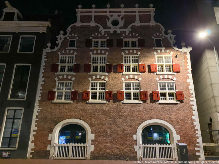 Night view of ornate brick building with stepped gable, white stone scrollwork, red shutters, and arched ground-floor openings.