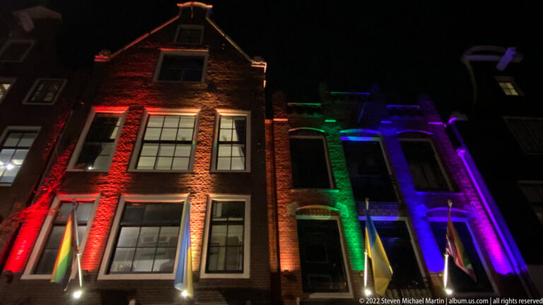 Narrow Amsterdam buildings illuminated at night with red, green, blue, and purple colored lights.