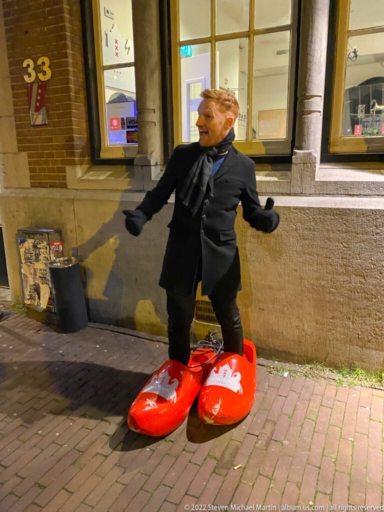 Smiling man wearing oversized bright red Dutch clogs on a brick-paved Amsterdam street.