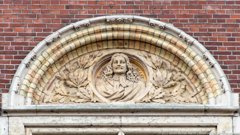 Round portrait bust in historical dress set within an arched, leaf-decorated surround on the Rijksmuseum façade.
