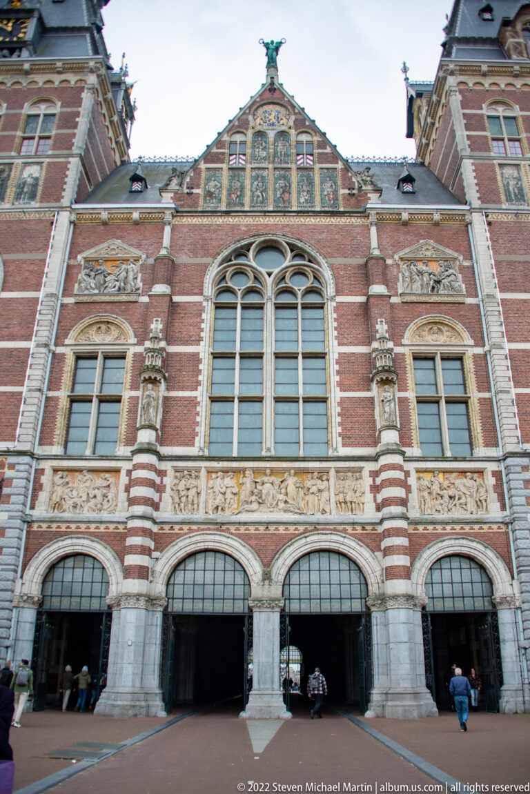 Frontal Rijksmuseum façade with broad passage arches, tall windows, decorative panels, and visitors below.