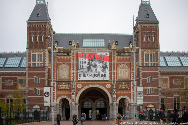 Wider view of the Rijksmuseum entrance, twin towers, central arch, exhibition banner, and pedestrians on the forecourt.