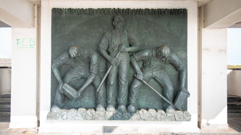 Bronze memorial relief with three male laborers, one standing and two crouching with tools.
