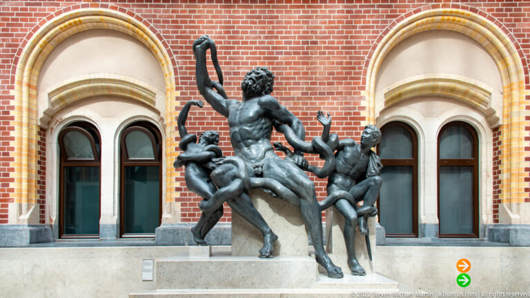 Bronze sculptural group with a tall nude central figure and several smaller figures before the Rijksmuseum wall.