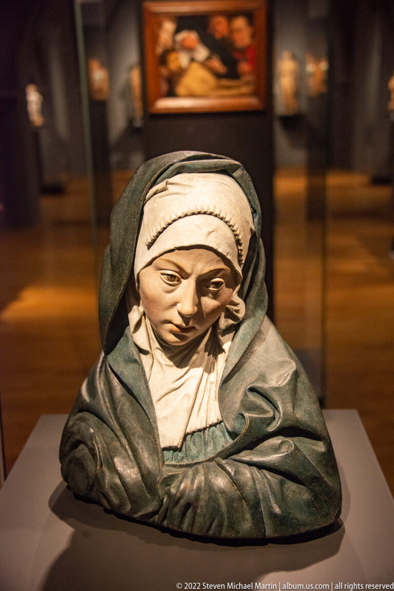Polychrome bust of a downcast woman wearing a white head covering and dark mantle on a museum plinth.