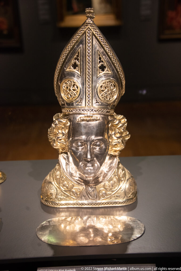Silver reliquary bust with a tall pierced mitre-like headdress, decorative rosettes, and broad metal base.
