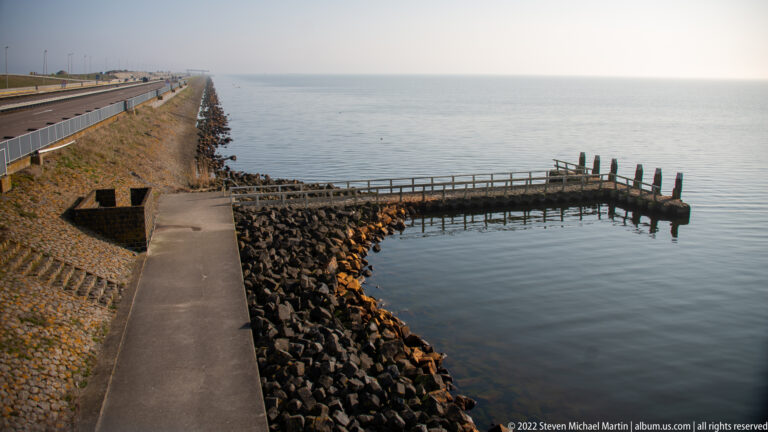 Paved path along a rock-armored shoreline leading to a narrow breakwater in calm water.